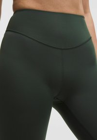 Magas derekú, sötétzöld leggings elasztikus anyagból, lapos varrásokkal és sima felülettel, diszkrét fényű, látható logók nélkül.