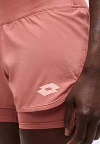 Tuvplāns cilvēkam, kurš valkā putekļaini rozā daudzslāņu sporta šortus ar gaiši rozā ģeometrisku logotipu sānos.