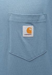 Světle modrá bavlněná košile s náprsní kapsou a bílou etiketou s oranžovým logem, prošitá dvojitým švem.