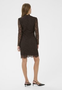 Rochie din dantelă maro, cu mâneci lungi, siluetă ajustată, margine cu unduiri și guler înalt. Asociată cu pantofi cu toc slingback negri.