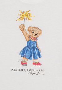 Bílý tričko s medvědem v modrých šatech, držícím žlutý pom-pom. Design zahrnuje logo a podpis ve spodní části.