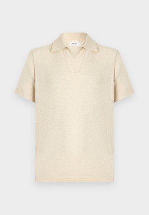 Uomo con capelli scuri e ricci che indossa una polo beige chiaro a maniche corte, pantaloni beige larghi, cintura nera e scarpe nere, in piedi con le mani nelle tasche.