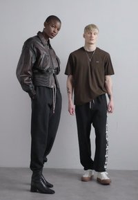 Zwei Modelle stehen zusammen. Das erste trägt eine braune, cropped Lederjacke, schwarze Hose und schwarze Stiefel mit Absatz. Das zweite trägt ein dunkelbraunes Shirt, schwarze Jogginghosen mit einem weißen Seitenaufdruck und weiße Sneakers.
