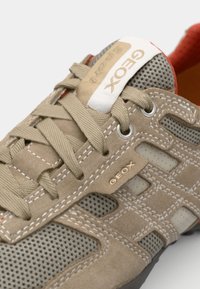 Beige semsket sko med mesh-paneler, snøring, gummisåle og rød innvendig. Har gullmerke på tungen og siden.