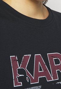 Černé bavlněné tričko s výrazným červeným a bílým grafickým textem s distressed efektem. Kulatý výstřih, krátké rukávky a jemná textura.