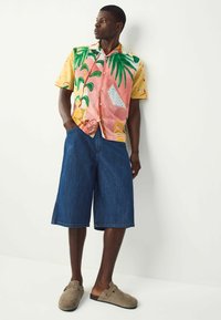 Camisa de botones de manga corta con estampado tropical en rosa y amarillo, combinada con pantalones cortos de denim hasta la rodilla y zapatos de tela marrón claro.