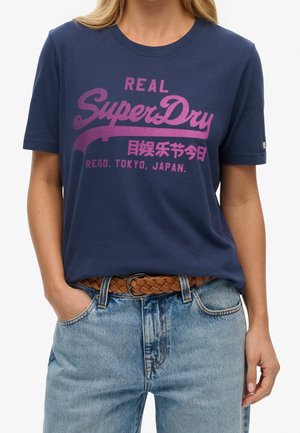 Kobieta nosząca granatowy t-shirt Superdry z różowym napisem i jasnoniebieskie jeansy z brązowym plecionym paskiem, ręce w kieszeniach.