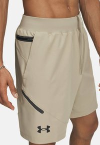 Beige sporta šorti ar gludu tekstūru, kas aprīkoti ar melnu rāvējslēdzēja kabatu, regulējamu jostasvietu un Under Armour logotipu uz apakšmalas.