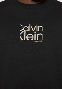 Černé bavlněné tričko s krémovým logem Calvin Klein v moderním písmu, umístěným na středu hrudi. Krátké rukávy a kulatý výstřih.