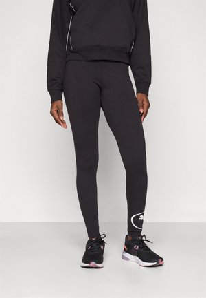 Leggings neri aderenti con una texture liscia, caratterizzati da un dettaglio del logo bianco sulla parte inferiore della gamba. Abbinati a scarpe da ginnastica nere.
