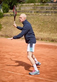 Tennista in una maglietta blu navy e pantaloni bianchi con dettagli turchesi, che tiene una racchetta, in piedi su un campo in terra battuta.