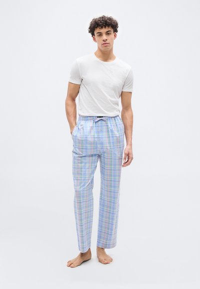 Νεαρός άντρας όρθιος ξυπόλυτος, φοράει λευκό κοντομάνικο μπλουζάκι και φαρδιά πιτζάμα σε παστέλ καρό σχέδιο με τα χέρια στις τσέπες.