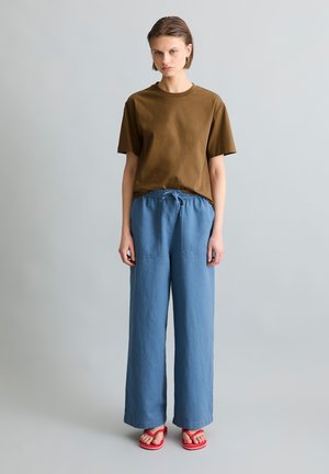 Jongere staand, met een bruin T-shirt met korte mouwen, losse blauwe broek met trekkoord en rode sandalen, effen lichtgrijze achtergrond.