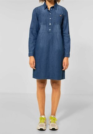 Vestido camisa de mezclilla, azul oscuro, de manga larga, con cuello, dos bolsillos en el pecho y botones en la parte delantera. Combinado con zapatillas claras.