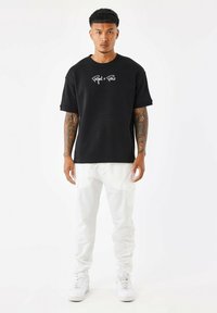 Camiseta de algodón negra con el texto en blanco "Project x Paris", combinada con pantalones cargo blancos. El modelo se encuentra de pie frente a un fondo plano.