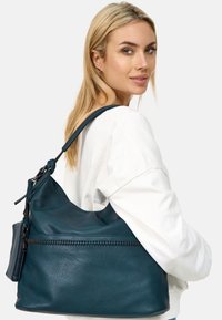 Teal leren handtas met een slouchy ontwerp, voorzien van een ritsvak en een gestructureerde afwerking, gedragen op de schouder met een lange schouderband.