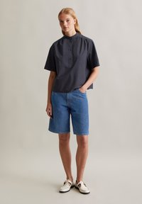 Camicia grigia scura a maniche corte con colletto alto, abbinata a pantaloni di denim blu corti fino al ginocchio. Il modello indossa scarpe bianche.