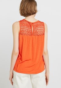 Pomarańczowy podkoszulek bez rękawów z koronkowym tyłem, z małym okienkiem i zapięciem na guzik, wykonany z miękkiego, lekkiego materiału.