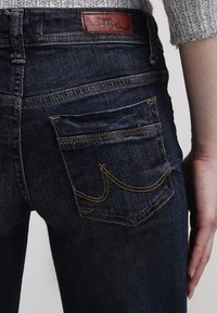 Jeans de mezclilla azul oscuro con costuras intrincadas en los bolsillos traseros y un parche de cuero con el logo de la marca en la cinturilla.