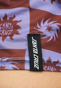 Κοντινό πλάνο μοβ και μαρόν καρό υφάσματος με ηλιοστάσεις και μαύρη ετικέτα με το όνομα "Santa Cruz" ραμμένη στην άκρη.