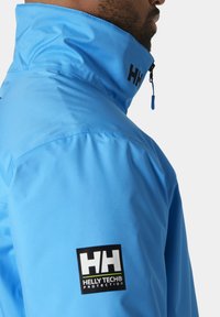Blauwe waterdichte jas met een hoge kraag, voorzien van een zwart logo-label en ritsaccent. De textuur is glad en glanzend.