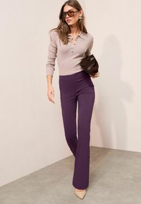 Lichtgrijze trui met een kraag en knoopdetails, gecombineerd met paarse flared pants en beige hoge hakken. Accessoires zijn onder meer een zonnebril en een bruine tas.
