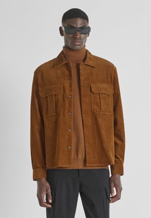 Bruine corduroy overhemdjas met twee borstzakken, knoopsluiting en een gladde textuur, gelaagd over een donkerder bruine coltrui.