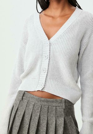 Cardigan blanc en tricot à col en V avec boutons, assorti à une jupe plissée grise à taille haute, laissant apparaître le ventre sur une personne aux cheveux longs tressés.