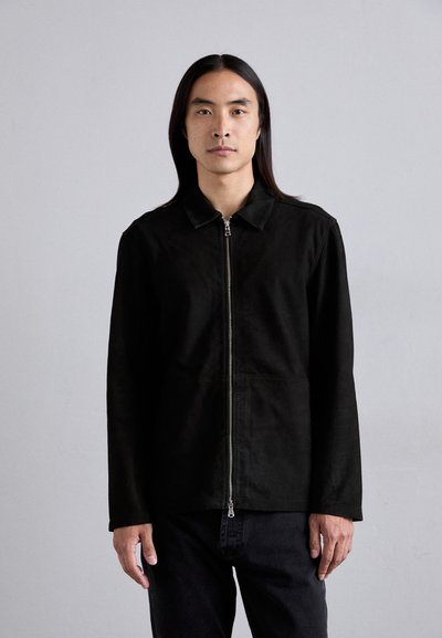 Sort zip-jakke med krave, lavet af blødt materiale, med lange ærmer og et minimalistisk design, båret over mørke bukser.