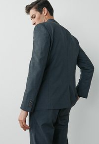 Mørkegrå skræddersyet blazer med en glat tekstur, enkelt ventil bagpå og fem knapdetaljer ved ærmerne. Bæres med hænderne på hoften.
