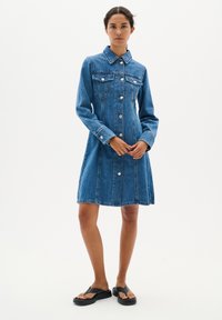 Robe en denim bleu moyen, avec un col, des manches longues, des poches avant et des fermetures à boutons argentés, présentant une silhouette à jupe évasée.