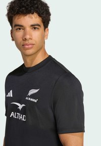 Sort atletisk trøje med korte ærmer, med en tekstureret stof, rund hals, "All Blacks" logo og hvid sponsortekst.