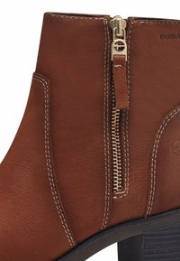 Brown leather ankle boot featuring a gold zipper, contrasting white stitching, and a low, chunky heel with a textured sole.