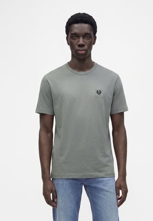 Junger Mann, der ein schlichtes graues T-Shirt mit kleinem Logo auf der Brust und hellblaue Jeans trägt, steht vor einem weißen Hintergrund.