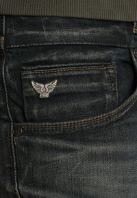 Los jeans de mezclilla oscura presentan un bolsillo frontal con un logo plateado alado, costuras naranjas y una textura suave.