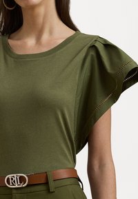 Maglietta verde in cotone con maniche corte e arricciate, dotata di tagli laterali, abbinata a una cintura marrone con una fibbia ovale argentata.