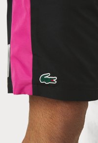 Melnas sporta bikses ar rozā tīkla sānu paneļiem un zaļo krokodila logotipa uzšuvumu. Tekstūrēta auduma konstrukcija ar brīvu piegulšanu.