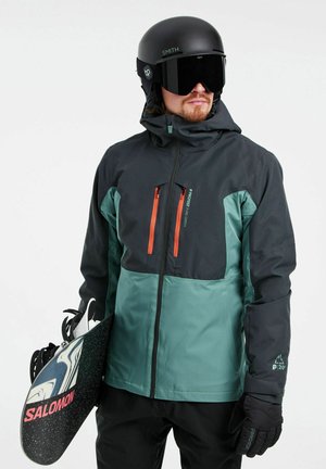 Schwarze und türkisfarbene Herren-Skijacke mit orangefarbenen Reißverschlüssen, strukturiertem Stoff, Kapuze und wasserdichtem Design. Snowboard in der rechten Hand gehalten.
