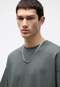 Maglietta grigia in cotone con scollo rotondo, adornata con una collana di catena argentata. Il modello ha capelli scuri e corti e un'espressione neutra.