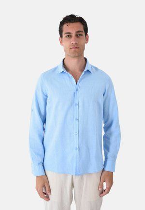 Uomo di fronte che indossa una camicia azzurro chiaro a maniche lunghe con bottoni e pantaloni beige su uno sfondo semplice.