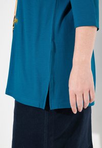 Türkises Baumwolloberteil mit abgerundetem Saum, kurzen Ärmeln und seitlichen Schlitzen. Verfügt über ein gesticktes Design nahe der Schulter. Kombiniert mit einem marineblauen Rock.