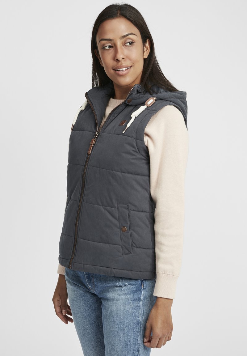 Gilet rembourré gris foncé avec capuche, doté d'une fermeture éclair à l'avant, de poches latérales et de cordons de serrage couleur crème. Porté sur un pull léger.