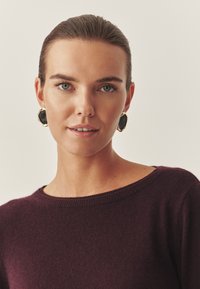 Bordo džemperis ar plašu kakla izgriezumu, kuru valkā modele ar īsiem matiem un lieliem melnbaltiem un zelta auskariem. Gluda tekstūra, minimālas detaļas.