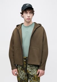 Καφέ φούτερ με φερμουάρ και τσέπες στο μπροστινό μέρος, φορεμένο πάνω από ανοιχτό πράσινο μπλουζάκι Nike. Συνδυάζεται με πράσινο και καφέ παντελόνι με σχέδια και μαύρο καπέλο.