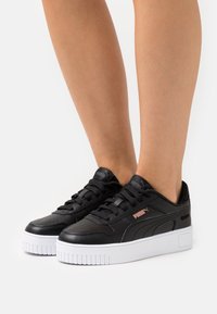 Personne portant des baskets Puma noires avec des semelles épaisses blanches, debout sur une surface blanche, montrant les jambes inférieures et les pieds.