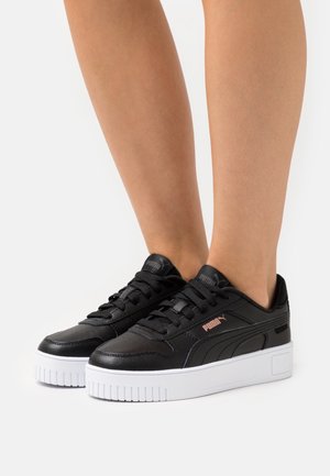 Personne portant des baskets Puma noires avec des semelles épaisses blanches, debout sur une surface blanche, montrant les jambes inférieures et les pieds.