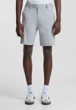 Shorts à carreaux gris avec une taille plate, des poches latérales et une fermeture à bouton. Portés avec des chaussettes de sport blanches et des baskets multicolores.