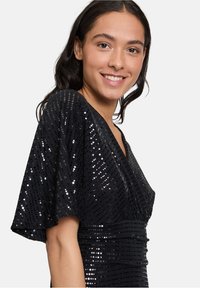 Robe noire ornée de sequins avec des manches courtes et flottantes, un décolleté en V et une taille cintrée, au surface texturée qui reflète la lumière.