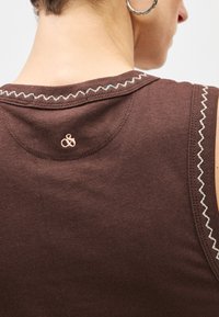 Καφέ αμάνικη μπλούζα με στρογγυλή λαιμόκοψη, με αντίθετη ραφή ζιγκ ζαγκ κατά μήκος της άκρης και μικρή κεντημένη λεπτομέρεια αμφερσάν.