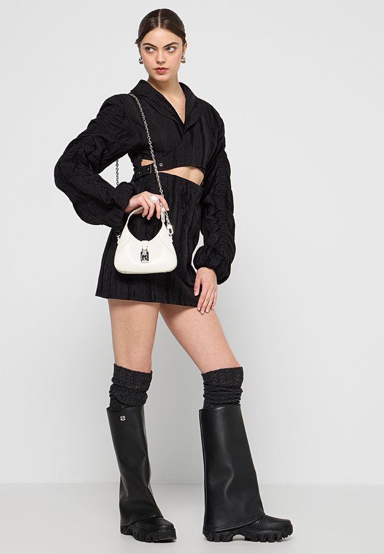 Top court noir texturé avec des manches ballons, mini-jupe noire, chaussettes grises et bottes en caoutchouc noires jusqu'aux genoux. Sac à main blanc avec une bandoulière en chaîne.